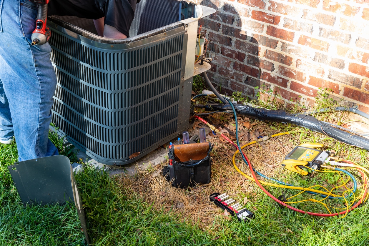 HVAC technician servicing an outdoor air conditioning unit beside a brick house with gauges, hoses, and tools on grass.