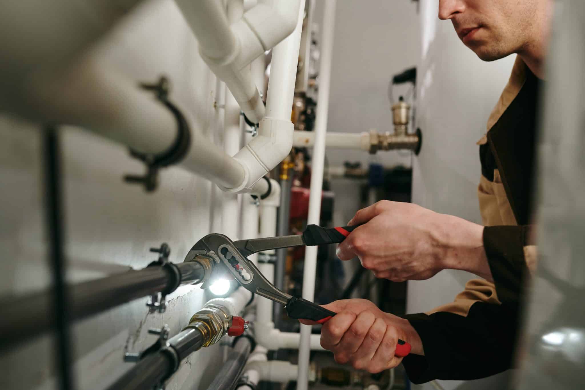 A plumber in overalls uses a pipe wrench to tighten a fitting on pipes in a mechanical/utility room.