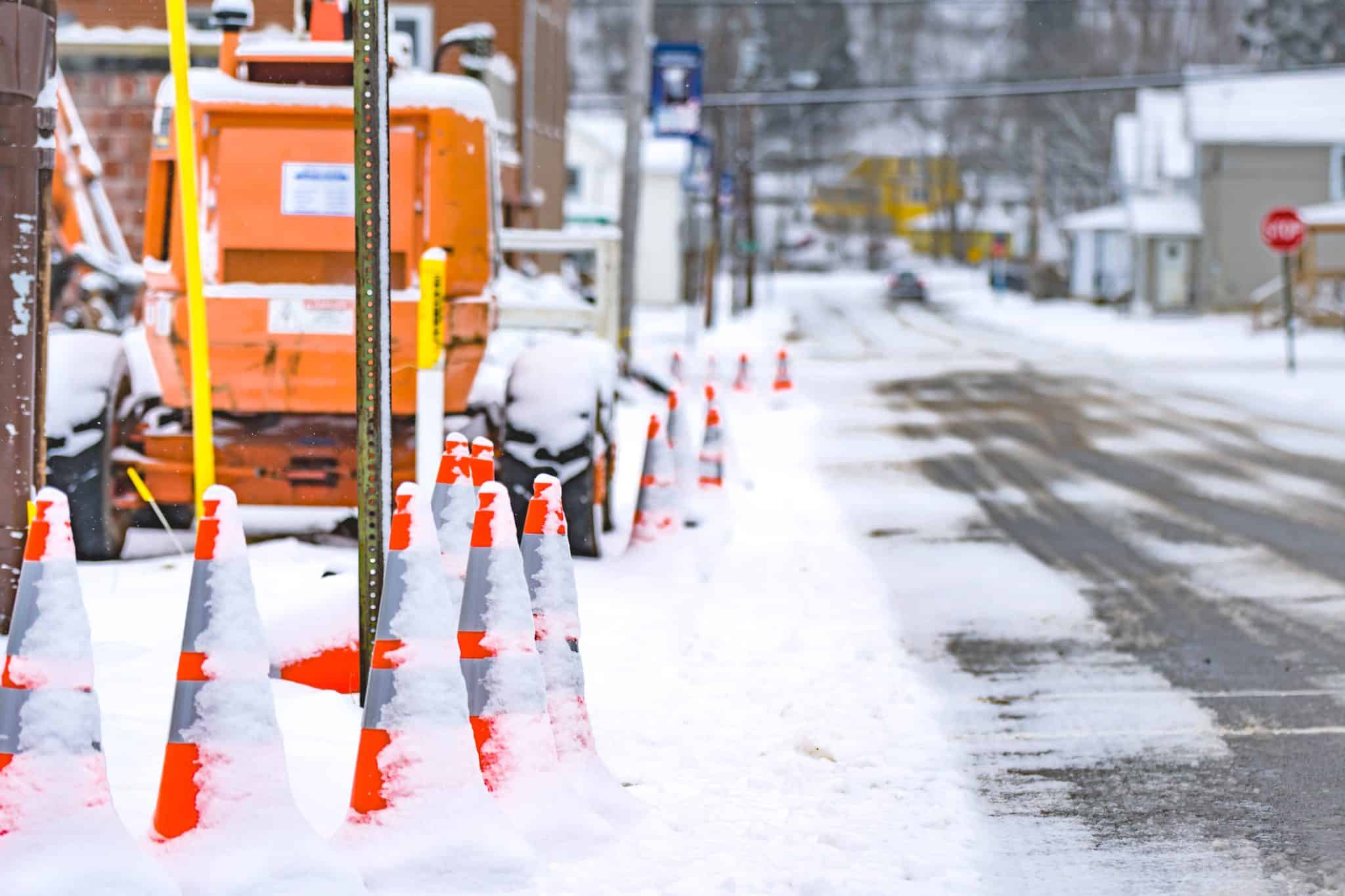 Snow-covered orange traffic cones line a quiet street beside a parked orange utility lift on a wintry day.